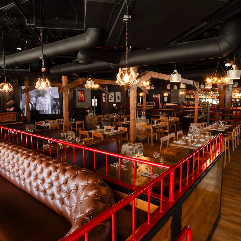 Rustic restaurant interior with leather booths, red railings, wood tables, exposed ducts, warm pendant lighting, and a stocked bar.
