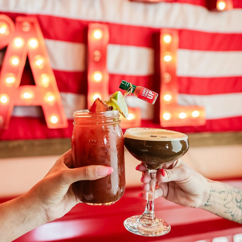 Hands hold a garnished red cocktail and a foamy dark drink against a glowing American flag backdrop.