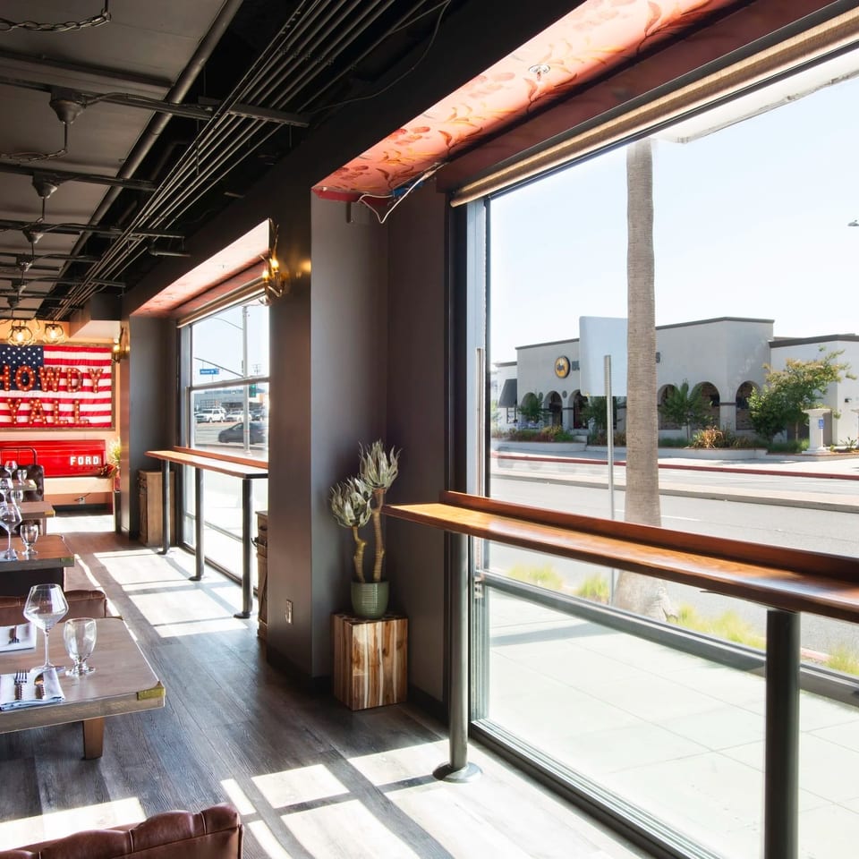 Bright restaurant dining room with set tables, leather seating, industrial lighting, and large windows overlooking a palm-lined street.