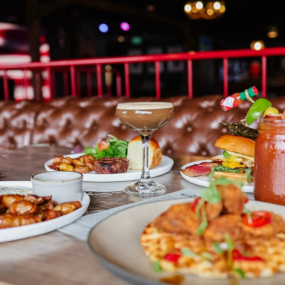 Restaurant table featuring assorted dishes, burgers, cocktails, and a Bloody Mary against a tufted leather booth and American flag backdrop.