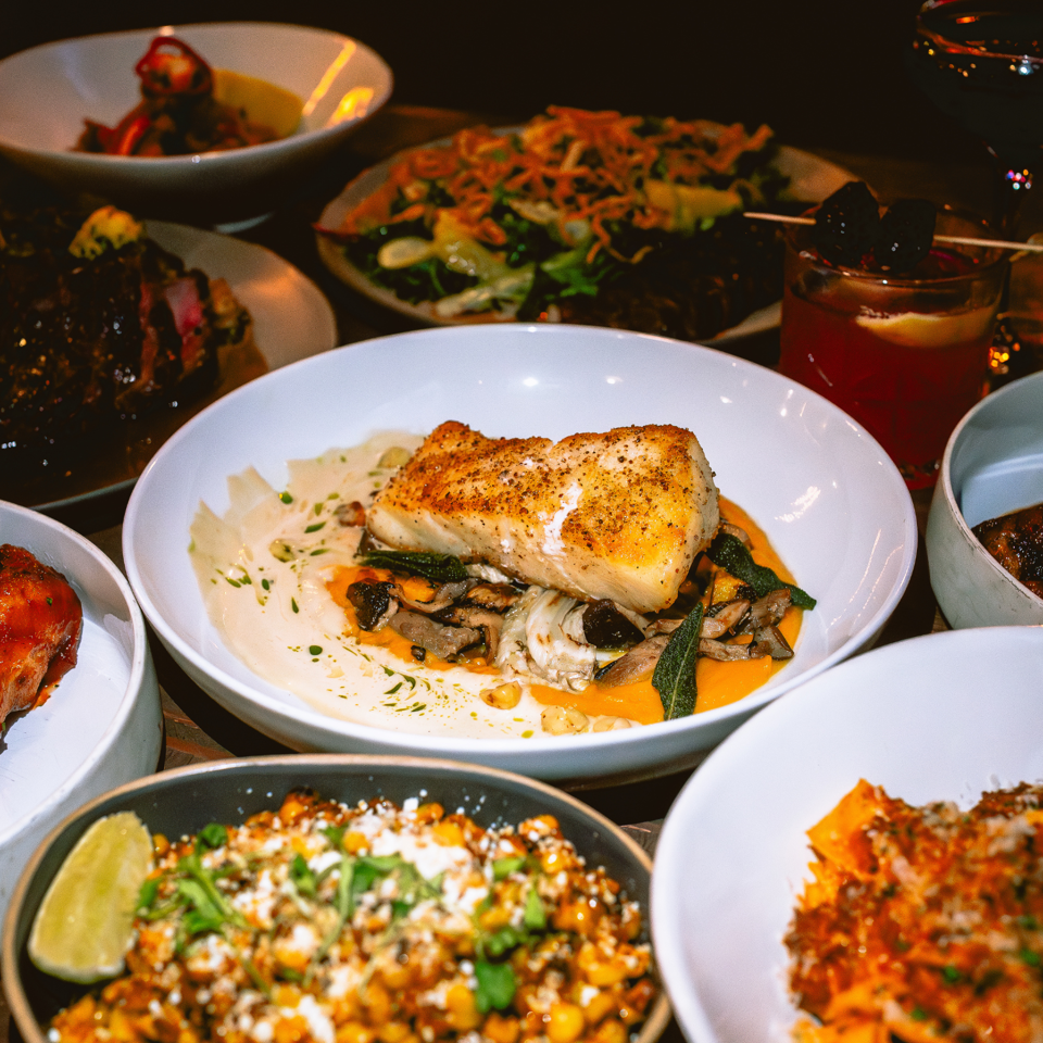 Dimly lit restaurant table featuring seared fish with mushrooms, colorful salads, corn, roasted vegetables, and cocktails.