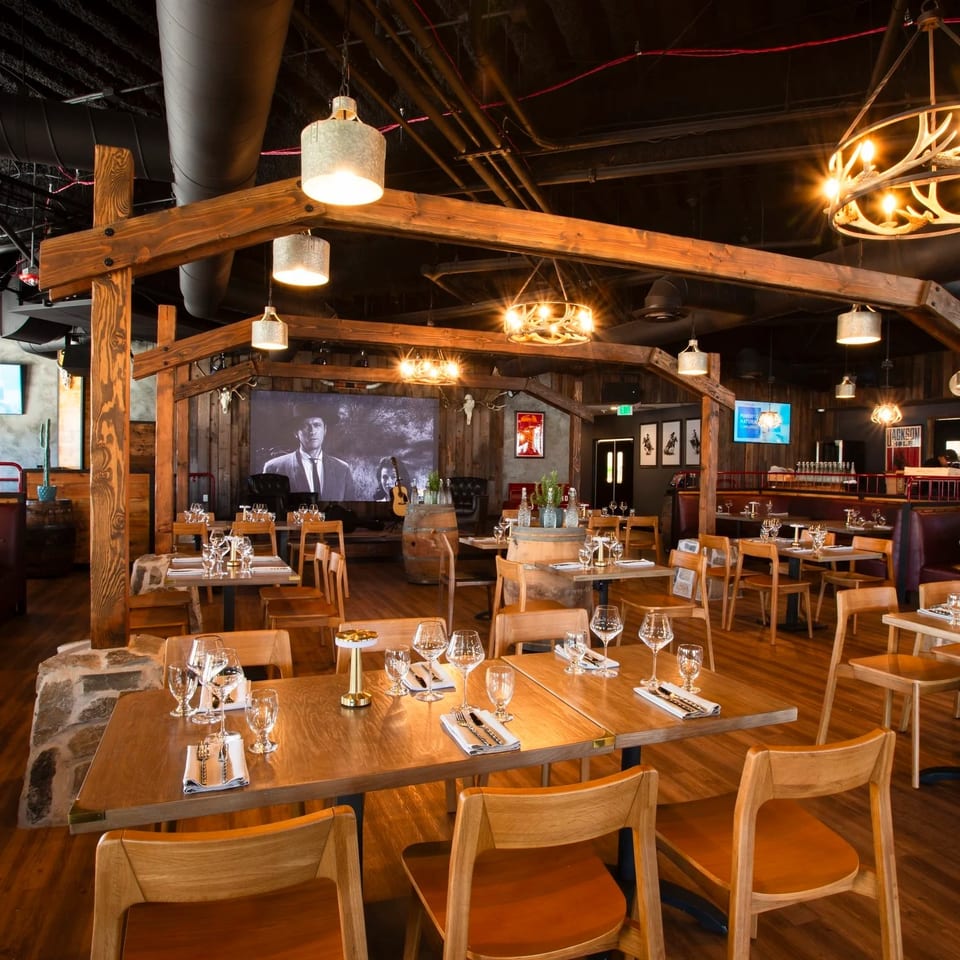 Rustic restaurant dining room with wooden tables, leather booths, exposed beams, antler chandeliers, and a central bar.