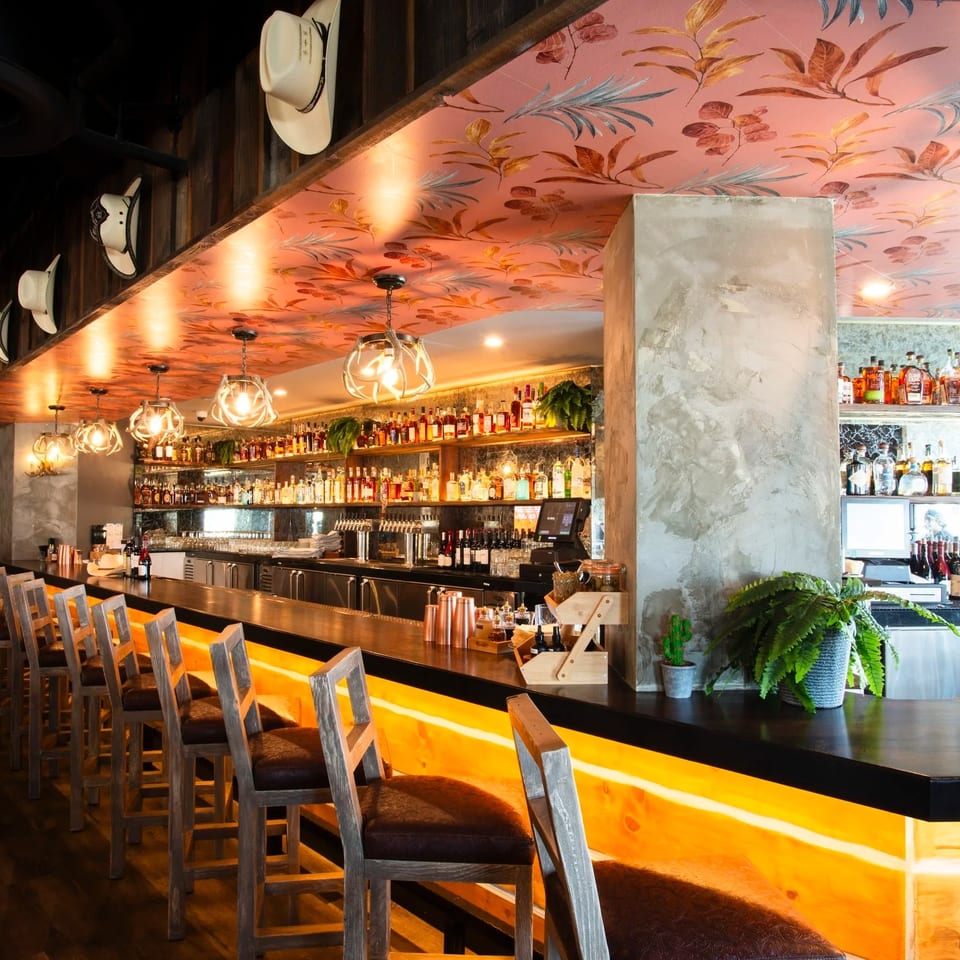 Warmly lit restaurant bar with rows of wooden stools, liquor shelves, floral ceiling, and antler-style pendant lights.