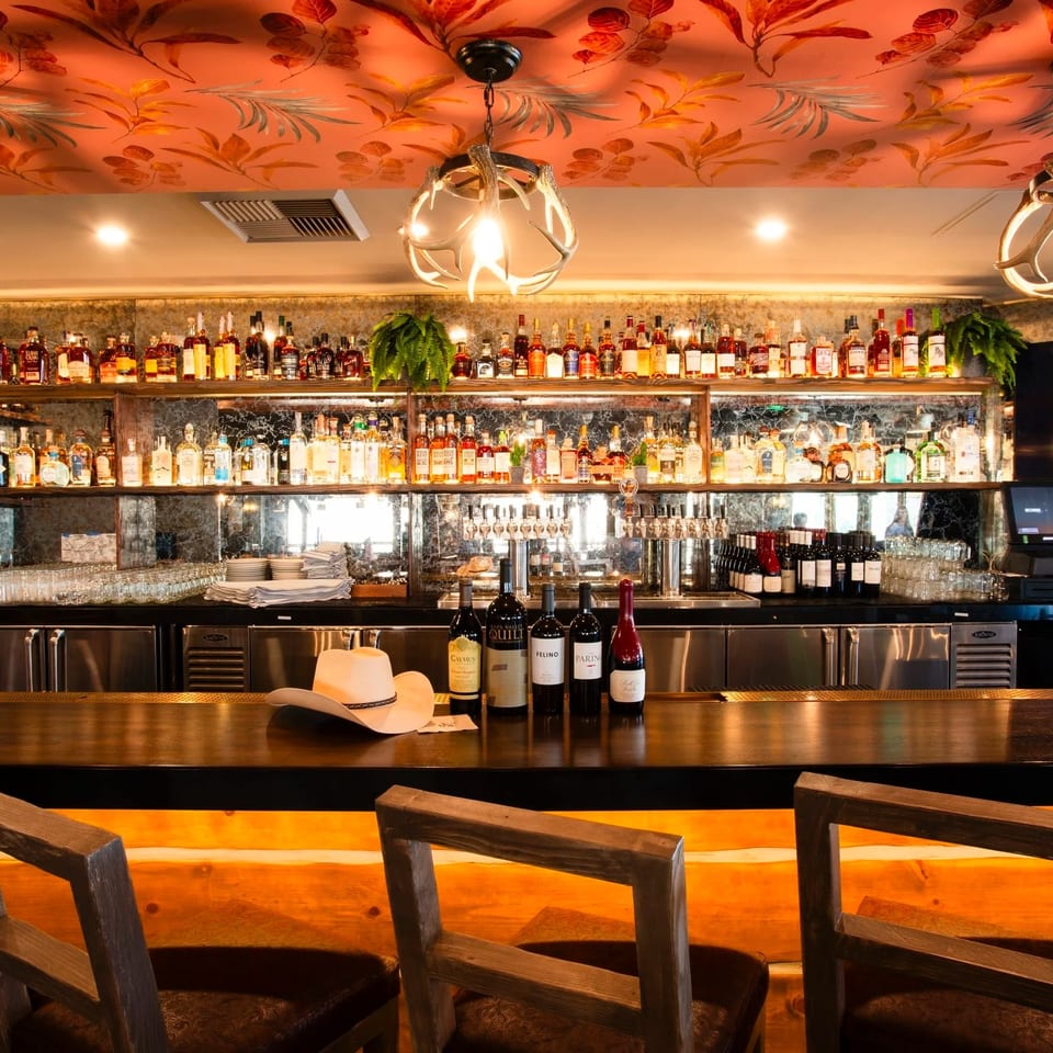 Warmly lit restaurant bar with shelves of liquor, hanging antler-style lights, wine bottles, television, and wooden seating.
