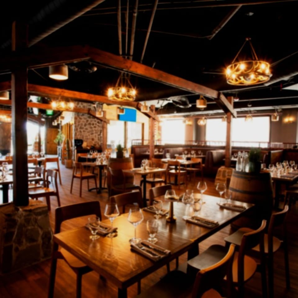 Warmly lit restaurant dining room with wooden tables, leather booths, exposed beams, wine glasses, and decorative chandeliers.