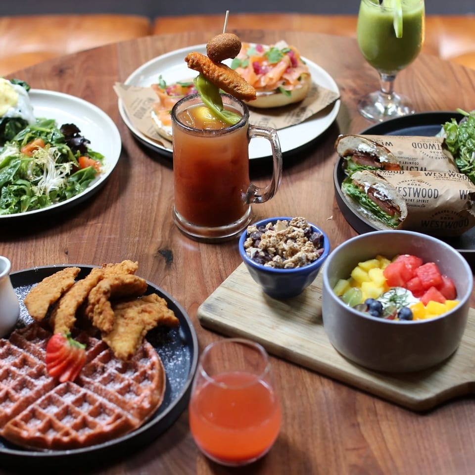 Restaurant table spread featuring salads, sandwiches, waffles with fried chicken, fruit, pastries, and colorful beverages.