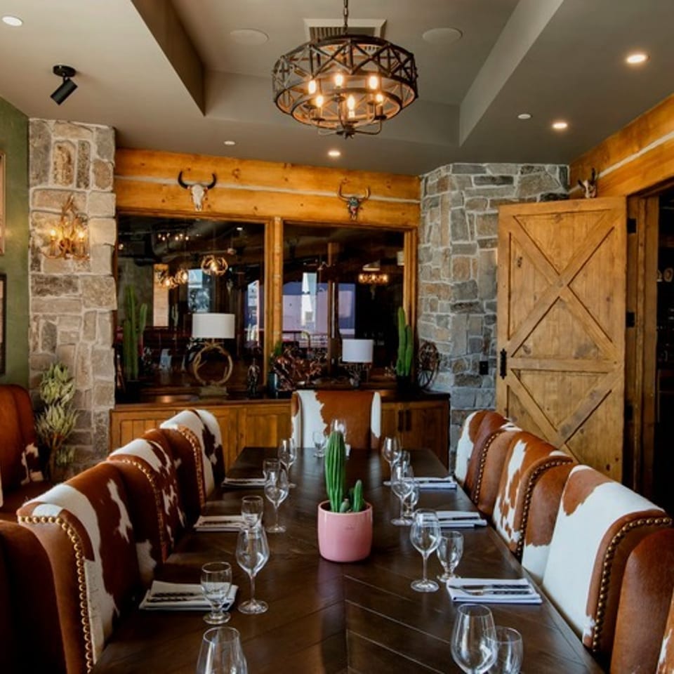 Rustic restaurant dining room with cowhide chairs, wooden tables, stone accents, Western artwork, and warm chandelier lighting.