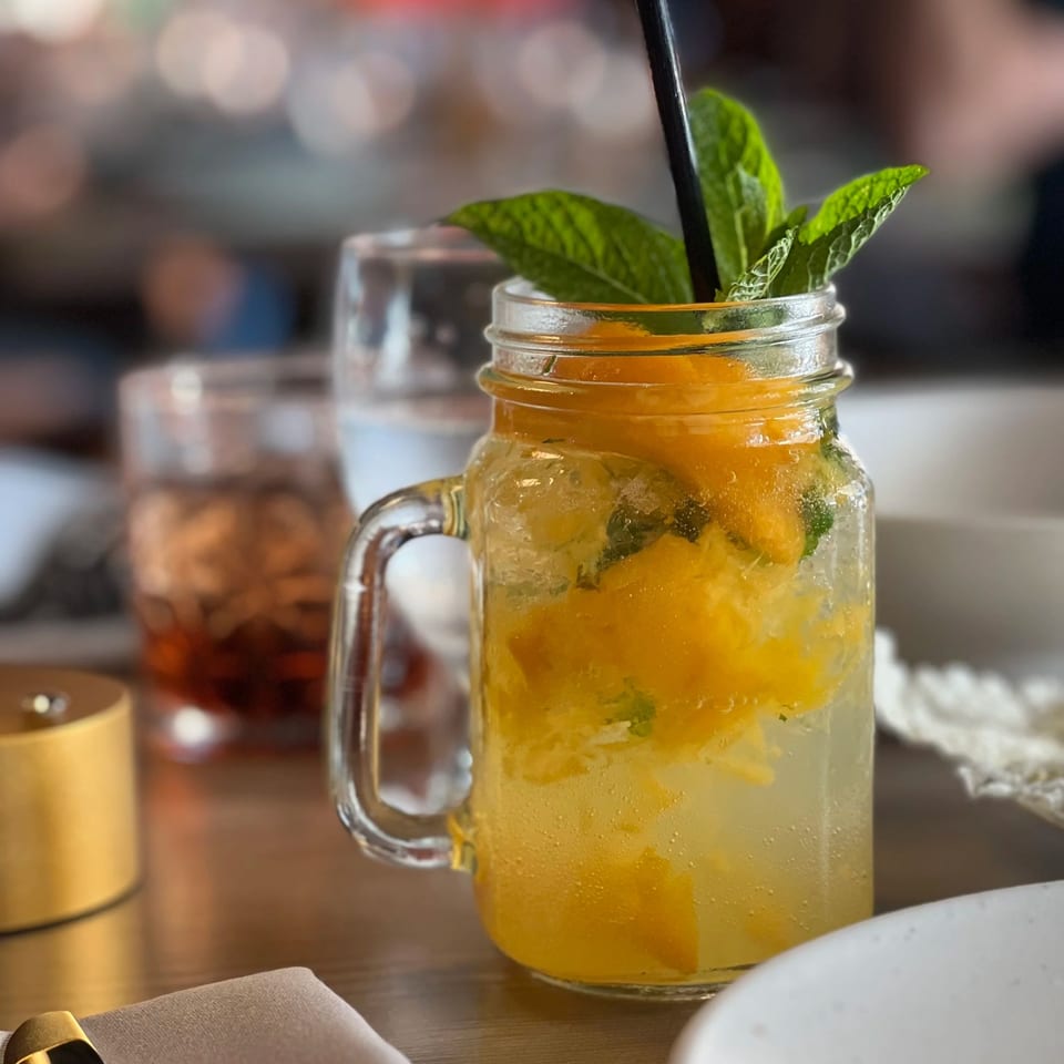A mason jar holds an orange iced drink with mint leaves and a black straw on a restaurant table.