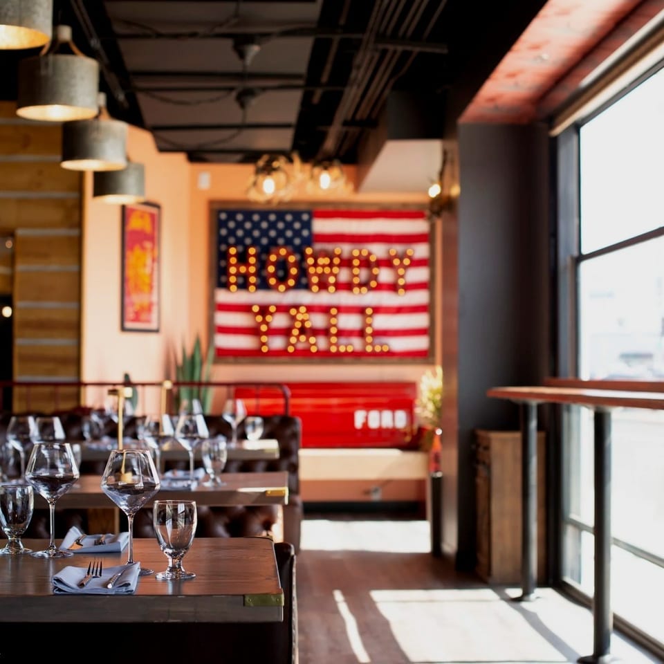 Warmly lit restaurant dining room with set tables, wine glasses, leather booths, large windows, and a “Howdy Y’all” American flag sign.