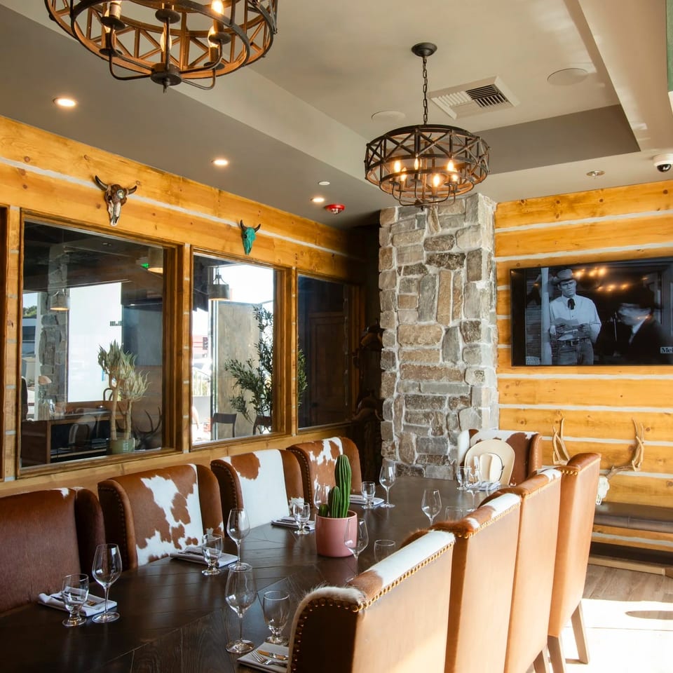 Rustic restaurant dining room with wood walls, stone columns, cowhide chairs, wagon-wheel chandeliers, cacti, and a television.
